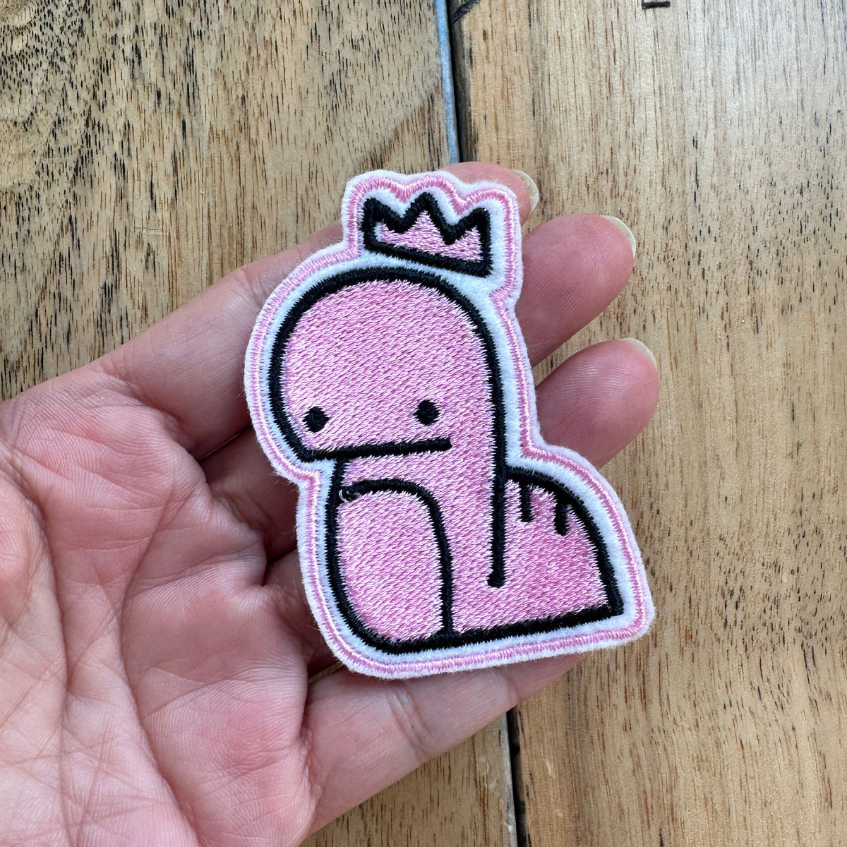 A hand holding a pink embroidered patch of a cartoon dinosaur wearing a crown, set against a wooden floor background.