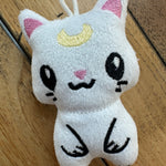 A small white plush cat keychain or charm with embroidered black facial features, pink inner ears, a yellow crescent moon, and a white hanging loop, photographed on a wooden surface.