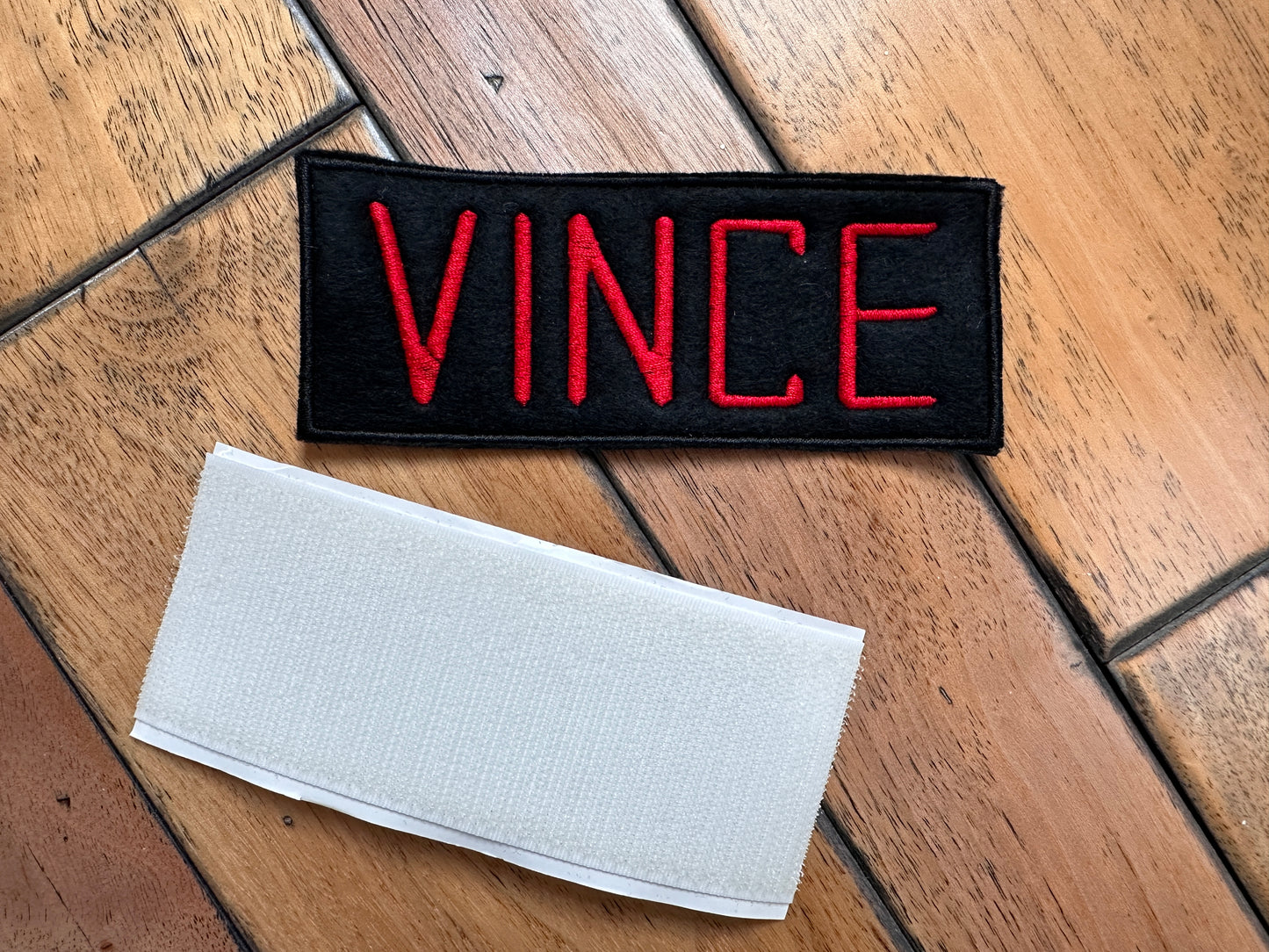 A black Ghost Buster style embroidered patch with the name 'VINCE' in red lettering, placed on a wooden surface above a white hook and loop strip.