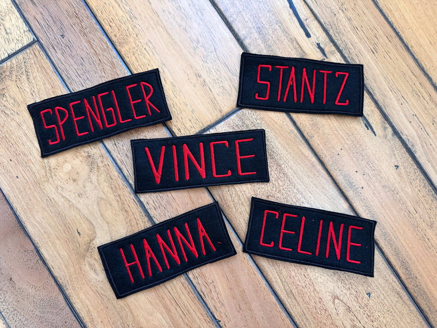 Five rectangular black embroidered name patches with red lettering in the style of Ghost Busters are arranged on a wooden floor. The names visible are SPENGLER, STANTZ, VINCE, HANNA, and CELINE.