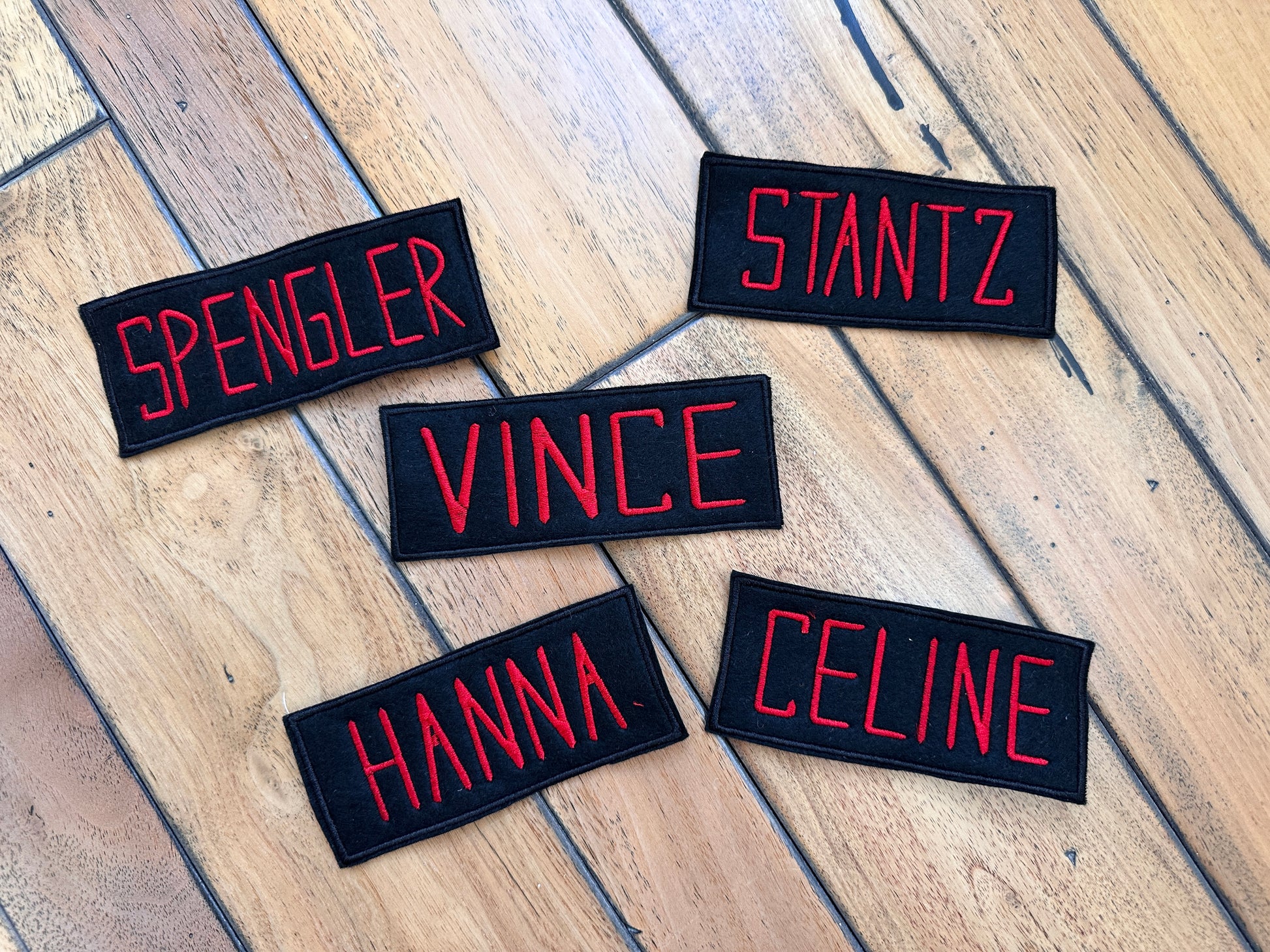 Five rectangular black embroidered name patches with red lettering in the style of Ghost Busters are arranged on a wooden floor. The names visible are SPENGLER, STANTZ, VINCE, HANNA, and CELINE.