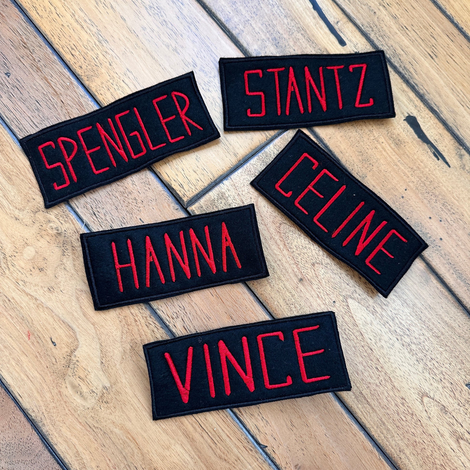 Five Bhost Busters embroidered name patches with red text names laid out on a wooden floor: SPENGLER, STANTZ, HANNA, CELINE, and VINCE.
