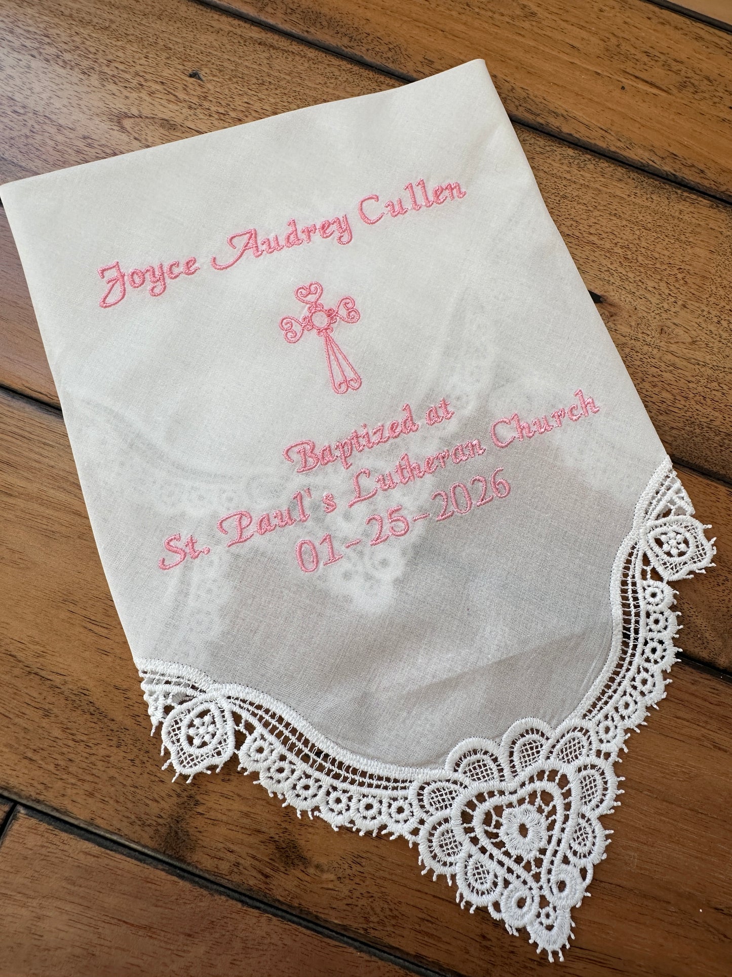 A square white lace handkerchief with pink embroidered text and a cross design, laid flat on a brown wooden surface. The text reads 'Joyce Audrey Cullen', 'Baptized at St.