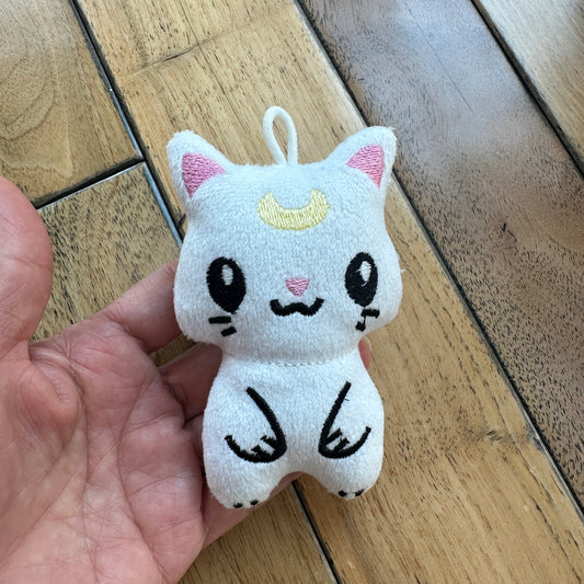 A small white plush cat charm with pink inner ears, a yellow crescent moon on its forehead, and black embroidered facial features and paws, held in a person's hand over a wooden floor.