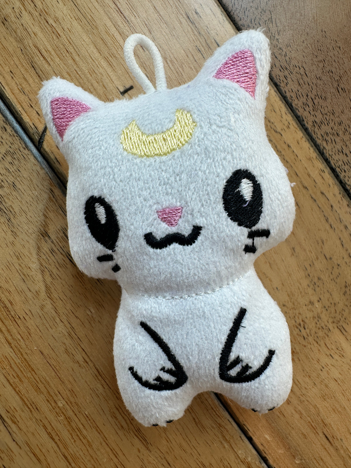 A small white plush cat keychain or charm with embroidered black facial features, pink inner ears, a yellow crescent moon, and a white hanging loop, photographed on a wooden surface.