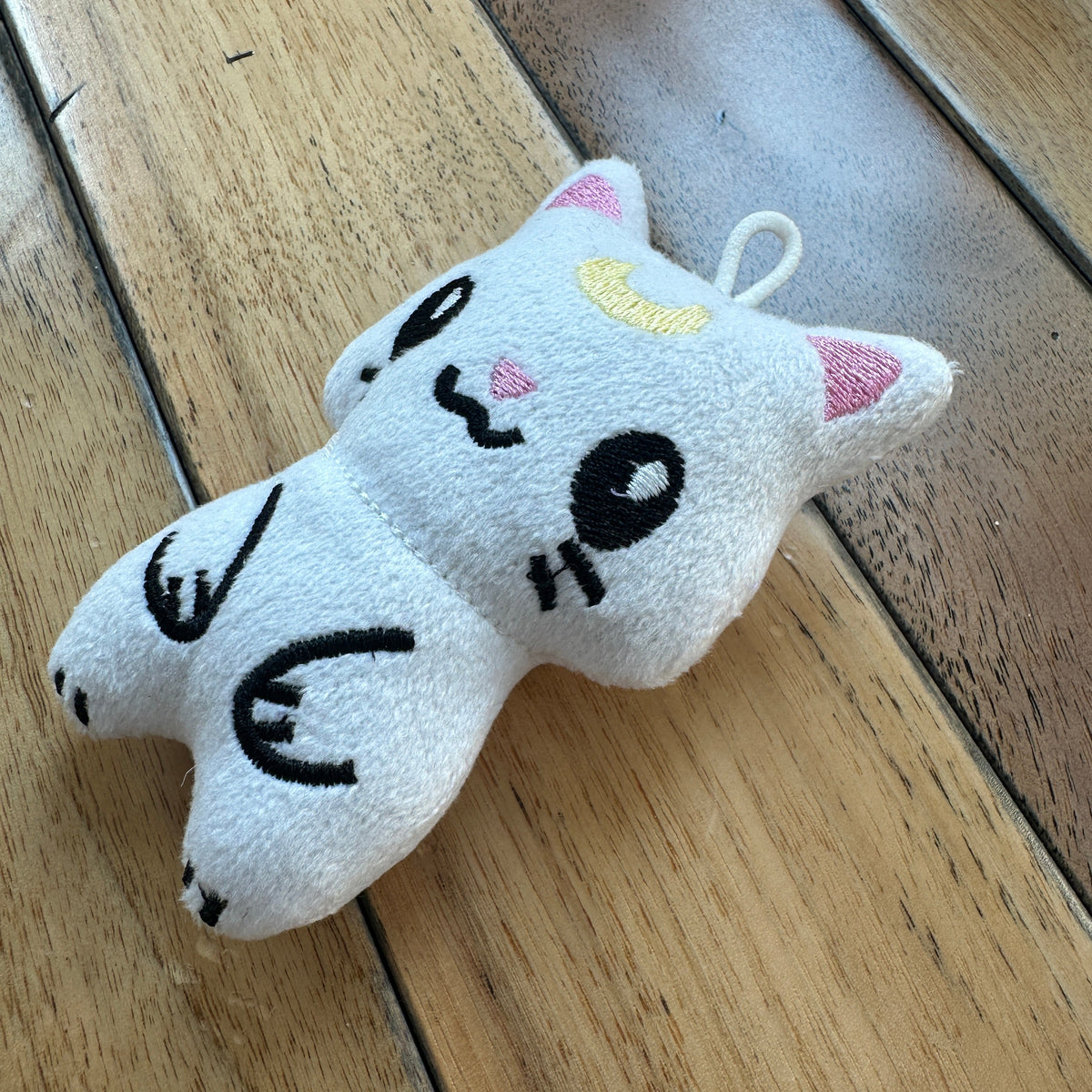 A white plush cat toy with embroidered black facial features, pink inner ears, and a small yellow crescent moon, lying on a wooden surface.
