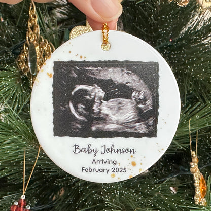 A round ceramic Christmas ornament featuring a baby ultrasound photo, personalized with the baby's family name and the year of birth, perfect for celebrating Christmasfor expecting parents and grandparents